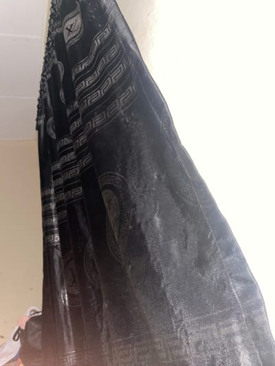 A close-up, low-angle shot captures a black, shiny, patterned curtain hanging against a white wall. The curtain exhibits a repeating geometric pattern and circular motifs, possibly a Greek key design and medallions. The fabric appears textured and reflects light, suggesting a velvet or similar material. The lower left corner shows a portion of a bag or bundle, with some pink and white fabric peeking out, resting on a surface. The overall setting appears to be indoors, possibly a residential space in Malete, Nigeria, indicated by the context of the request.