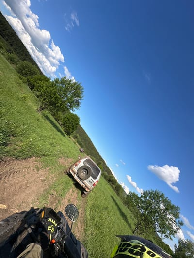 The image depicts a landscape with a dirt track running through a grassy field in the direction of a forest, possibly in the vicinity of Пепеляева, Russia. A silver SUV, with a spare tire mounted on the back, appears to be parked on the track ahead. In the foreground, part of an all-terrain vehicle (ATV) is visible, including a mirror and the rider's hand wearing a glove with "FOX" branding. The sky is blue with scattered clouds. The image is angled as if taken from the perspective of the ATV rider. The scene is outdoors, and the environment suggests an off-road or rural setting.