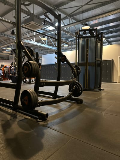 This image depicts the interior of a gym, likely located in Alès, France. The scene is dominated by various weightlifting equipment, including a prominent squat rack with weight plates and a cable machine in the background. The gym floor is made of dark, textured rubber tiles. Overhead, exposed ceiling structures, lighting fixtures, and an air conditioning unit are visible. Lockers are lined up against a wall in the mid-ground. Two individuals are partially visible in the background, appearing to be engaged in a workout or observing the equipment. The overall atmosphere suggests a modern and functional fitness facility.