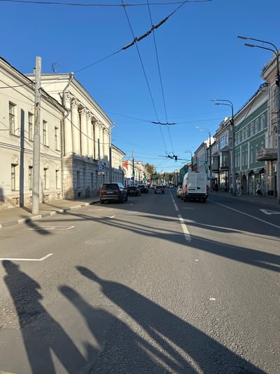 The image shows a street scene in the city of Feklino, Russia, on a clear day. The foreground is dominated by the asphalt road, with shadows of people cast across it. In the middle ground, several cars are driving on the road, including a white van and a dark SUV. On the left side of the road, there is a building with classical architecture, featuring columns and ornate detailing. A blue parking sign is visible on this building. On the right side of the road, a row of buildings with shops on the ground floor line the street. The sky is a bright, clear blue, with overhead wires for what appear to be trolleybuses crisscrossing the scene. The overall environment suggests an urban setting with active street life.