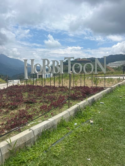 The image depicts a large, white, three-dimensional sign spelling out "HOBBITOON" in uppercase letters. The sign is positioned in an outdoor setting with lush green grass and a border of red-leafed plants in the foreground. Behind the sign, a building with a distinctive roofline is visible on a hillside, surrounded by trees and mountains. The sky above is partly cloudy with a mix of blue sky and white clouds. In the background, there appears to be some sort of terraced agricultural or horticultural development on the slopes of the hills. The overall scene suggests a tourist attraction or themed park. The location is identified as Kampung Kerawat, Malaysia. There are no people visible in the image. The scene is clean except for a few pieces of litter on the grass.