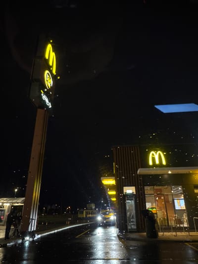 In the city of Laagri alevik, Estonia, at night, a McDonald's restaurant is visible. A tall signpost with illuminated McDonald's arches stands to the left of the frame. The McDonald's building itself is lit, with a neon arch visible on its facade. The ground in front of the restaurant is wet, reflecting the lights and suggesting recent rain. A few scattered raindrops are visible on the lens of the camera. In the distance, a yellow vehicle, possibly a bus or tram, is seen approaching with its headlights on. Two individuals are standing near the base of the signpost on the left side of the image. The overall scene depicts a typical urban nightscape at a fast-food establishment.