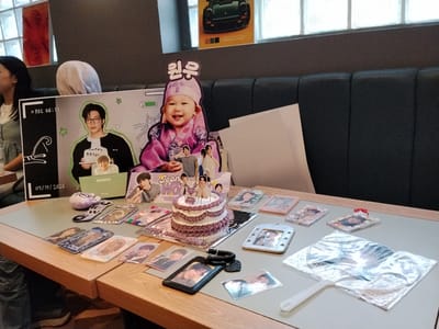 A birthday celebration is underway at a restaurant table. A young child, dressed in traditional purple Korean attire and a matching hat, is featured prominently on a large cardboard cutout. The child is smiling and appears to be the centerpiece of the event. Surrounding the cutout are various merchandise items, including photo cards, keychains, and a cake decorated with purple frosting. The table is set with these items, suggesting a fan gathering or a birthday party for a fan of the person depicted on the cutout. The background shows a booth seating area typical of a restaurant, with a dark gray upholstered bench. The overall atmosphere is festive and celebratory. The presence of Korean cultural elements suggests the individual being celebrated might have Korean heritage or be a fan of Korean culture.
