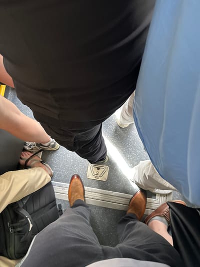 The image shows a close-up, bird's-eye view of the feet of three people standing on the floor of a public transportation vehicle, likely a bus. The vehicle is in the town of Griante, Italy, located at latitude 45.985 and longitude 9.237. The person in the center of the image is wearing brown leather shoes. The person on the left is wearing brown leather sandals. The person on the right is wearing brown leather shoes. The floor is gray with a metal strip running diagonally across the center. There is a rectangular metal panel with a circular, raised design at the bottom of the center of the image. The only visible feature of the vehicle is a bright sliver of sunlight reflecting on the floor. The people in the image are wearing casual clothing. The person in the center is wearing dark pants and a white shirt. The person on the left is wearing brown pants. The person on the right is wearing blue jeans and a blue shirt. All three people appear to be casually standing in the vehicle, perhaps waiting to reach their destination.