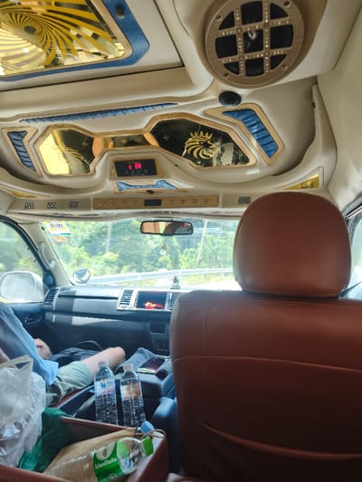 The image captures the interior of a customized passenger vehicle, likely a van or bus, in transit through Ban Wang Pra Chan, Thailand. The viewpoint is from a rear seat, looking forward towards the front cabin. The vehicle's ceiling is elaborately decorated with cream-colored upholstery, gold-colored trim, and blue textured accents. Overhead features include gold-mirrored panels, one displaying a crowned lion emblem, a large spiral design in gold, and a central digital display showing "88" in red, along with a clock icon. A circular speaker grille is visible in the upper right.
In the immediate foreground, a brown leather headrest occupies the right side of the frame. Forward of this, the lower torso and legs of a passenger, wearing light-colored shorts, are visible in the front passenger seat. Between the front seats, on a central console, two clear plastic bottles of Singha R-O drinking water stand upright, next to a dark smartphone or tablet resting on a grey cloth. A dark bag is positioned on the floor or console area in front of the passenger. In the lower left, a white plastic bag and a green-capped "TIP" brand water bottle are contained within a brown receptacle.
Through the front windshield, a multi-lane road with a white metal guardrail is visible, bordered by dense green foliage and trees under a bright daytime sky. A small, dark figurine, possibly a Buddha statue, is present on the dashboard. A rearview mirror hangs centrally, with a small decorative item suspended from it. The dashboard appears standard for a commercial van.