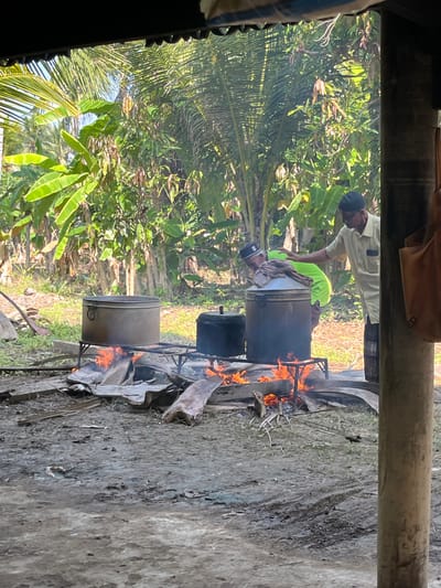 This image depicts an outdoor cooking scene in the village of Peurupok, Indonesia. Two men are tending to large metal pots set over open fires, which are fueled by wood and other debris. The area appears to be rustic, with a dirt ground surface and lush tropical vegetation, including banana trees and palm trees, in the background. One man, wearing a light-colored shirt and a black cap, is standing and interacting with the other man. The second man, dressed in a green shirt and a black cap, is leaning over one of the large pots, seemingly attending to the cooking process. A portion of a wooden structure with a dark roof is visible in the foreground, suggesting a covered area or shelter. The overall impression is one of traditional food preparation in a rural, tropical environment.