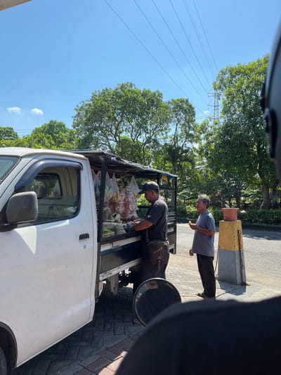 This image depicts a street scene in Pagerwojo, Indonesia, under a clear blue sky with scattered clouds and visible power lines overhead. The focal point is a white truck modified to serve as a mobile food stall. The truck's bed is covered with a tarp, displaying various food items packaged in plastic bags and hung from the frame.
Two men are present near the stall. One, wearing a dark t-shirt, a baseball cap, and a crossbody bag, appears to be tending to the food items. The other man, wearing a grey t-shirt and dark pants, stands to the right, holding a small food item in his hand, suggesting he is a customer.
The ground is paved with interlocking bricks. To the right of the men, a yellow and grey concrete post with chains is visible, and further back, lush green trees and vegetation line the area. The overall impression is a typical roadside scene in a tropical climate where mobile vendors are common.