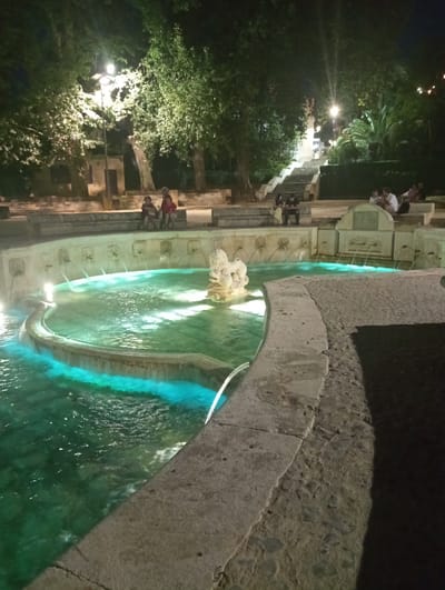 The image shows the Fountain of the Lions (Fuente de los Leones) in Priego de Córdoba, Spain, at night. The fountain is centrally located, with a large, multi-tiered pool of water illuminated with a vibrant turquoise glow. Several spouts emit streams of water into the main pool, and a decorative stone sculpture of a lion figure sits within the fountain's upper section. The fountain is surrounded by stone pathways and tiered seating areas. Several people are visible in the background, seated on the stone benches and steps. Some are engaged in conversation, while others appear to be looking at their phones. The scene is further enhanced by overhead lighting from streetlamps, casting shadows and highlighting the architectural details of the fountain and its surroundings, including large trees framing the upper part of the image.