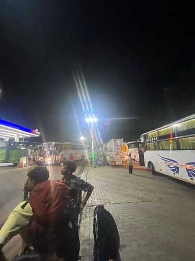 The image depicts an outdoor scene at night, likely a gas station in Kajheri, India, indicated by signage. A white bus with "INDO CANADA" written on its side is prominently featured, along with other vehicles including a green bus and a tanker truck with "HP" branding. Several individuals are present, some appearing to be passengers waiting by the buses. One person is standing near the tanker truck. The ground is paved with bricks, and bright lights illuminate the area. The overall atmosphere suggests a busy transit point at night.
