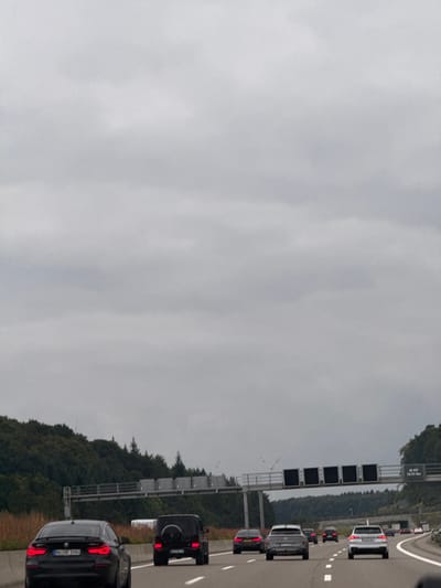 The image displays a multi-lane segment of the German Autobahn 8 (A8) near Dornstadt, Germany, under a uniformly overcast sky. The roadway features multiple lanes of traffic, with vehicles proceeding in the same direction as the viewer. In the immediate foreground, a black BMW 4 Series Gran Coupé, bearing the German license plate `HD HV 9556`, occupies the left lane. Ahead of it, a black Mercedes-Benz G-Class SUV is positioned in the middle lane. Further down the road, several additional vehicles are observed, including a gray Audi SUV and a white SUV in the right lane, and a dark sedan in the left lane. An overhead gantry spans the highway, supporting multiple black, unlit variable message signs and a fixed sign on the right side displaying "A8 - PÖL..." and a kilometer marker "136,777 km". Dense forest, composed of both deciduous and coniferous trees, lines the elevated terrain flanking both sides of the highway. The sky is entirely covered by a thick layer of gray clouds. No individuals are visible.