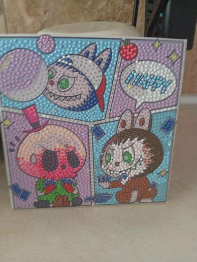 The image shows a diamond painting artwork featuring cartoon characters. The artwork is divided into four panels, mimicking a comic strip. In the top left panel, a character with large green eyes and a red mouth is blowing a pink bubble. The top right panel displays a rabbit-like character with large green eyes and a speech bubble containing the word "HAPPY". The bottom left panel features a skull-like character wearing a pink top hat and holding playing cards. The bottom right panel depicts another rabbit-like character with green eyes, a mischievous grin, and playing cards in its hands. The background of each panel is a textured pattern of small dots in various pastel colors, including blue, pink, and white. Small yellow star shapes are also scattered across some panels. Playing cards are depicted scattered around the characters in the bottom panels. The artwork appears to be placed on a flat surface, possibly a table, with a blurred background that suggests an indoor setting. The lighting appears to be natural and soft, casting a slight shadow. There are no visible landmarks or elements in the image that can be used to determine its location in Chigatay, Uzbekistan.
