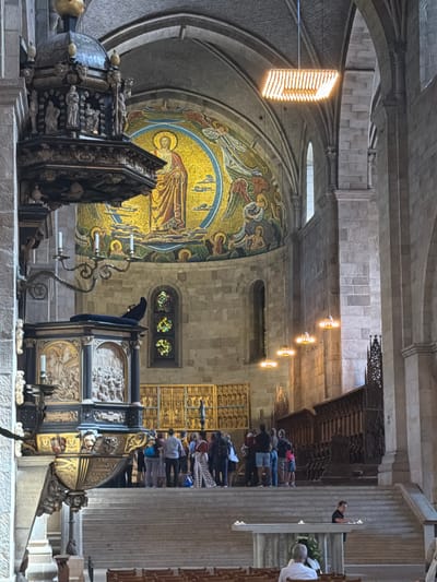 The image shows the interior of Lund Cathedral in Lund, Sweden. In the foreground, a stone altar with several lit candles is visible, with two individuals, one seated and one standing, in front of it. A broad set of stone stairs leads up to a platform where a group of tourists are gathered, some looking at a mosaic on the wall. To the left, a detailed pulpit is prominently featured, adorned with carvings and decorative elements. Above the pulpit, a large mosaic depicting a central figure with a halo and surrounding figures is displayed on the curved wall of the apse. Hanging from the ceiling are several contemporary light fixtures, contrasting with the historical architecture of the cathedral. The cathedral's stone walls and arched ceilings are characteristic of Romanesque architecture.