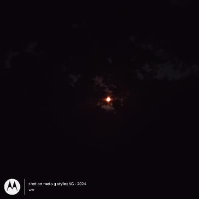 The image shows a full moon shining brightly in the night sky. The moon appears to be partially obscured by clouds. The image was taken in Happy Valley, Oregon, United States, at the coordinates 45.443, -122.552. The image was captured with a Motorola Stylus 5G phone in 2024. No people or other objects are visible in the image.
