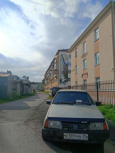 The image depicts a street scene in Shingarskie Isady, Russia. In the foreground, a light-colored car is parked on the right side of a paved road. The car has a license plate that reads "B 389MB 35". To the right of the car, a black metal fence runs along the edge of the property. Beyond the fence, a multi-story building with light-colored walls and balconies is visible. A large mural featuring a detailed painting of a samovar and other objects is present on the side of an adjacent building to the left of the multi-story one. The street continues into the distance, lined with more buildings and some trees. The sky above is partly cloudy with a mix of blue sky and white clouds.