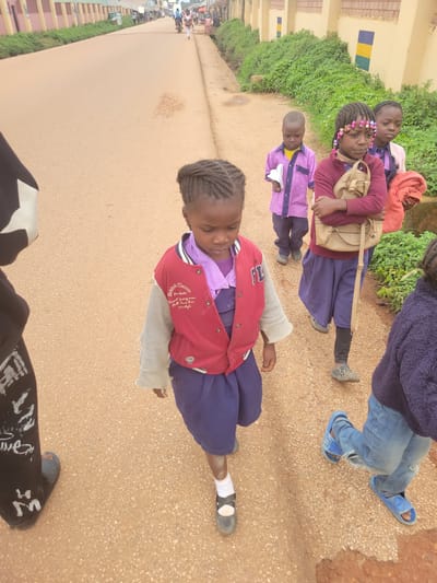 The image shows a group of children walking on a dusty road in Tangu, Nigeria. The children are wearing school uniforms, and some of them are carrying bags. The weather appears to be sunny, and the time of day is likely daytime, given the lighting. The children seem to be in good spirits, with some of them smiling.

In the foreground, a young girl in a red and purple jacket is looking at the camera. She has braids and is wearing a purple shirt. Her jacket has some text on it, but it is difficult to read. Behind her, other children are walking. One girl in a maroon jacket and pink hoodie is wearing a beige bag. Another child in a purple uniform is holding a piece of paper. Further back, more children are visible, as well as adults and motorcycles, suggesting a busy street scene.

The setting appears to be a residential or school area, with buildings and lush greenery lining the road. The road itself is unpaved and dusty, characteristic of many roads in rural Nigeria. The overall impression is one of everyday life in a Nigerian community, with children on their way to or from school.