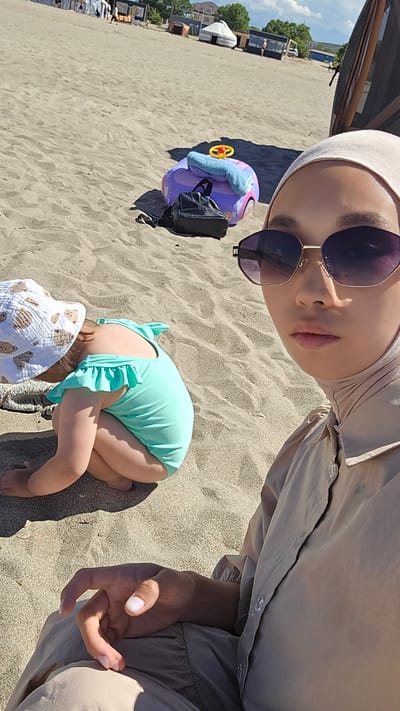 This is a vertically oriented, outdoor, eye-level selfie taken on a sunny day at a beach. The foreground features the face of a woman wearing a light pink hijab and large, dark-rimmed sunglasses. She is looking towards the camera. Next to her, her hand is raised, with the index finger and thumb touching, forming a relaxed gesture.
In the mid-ground, a young child is sitting on the sand. The child is wearing a mint green swimsuit with ruffled details and a white bucket hat adorned with teddy bear illustrations. Behind the child, a purple toy car with a blue towel and a yellow steering wheel is casting a shadow on the sand. A black bag is placed on top of the toy.
The background shows a sandy beach with rows of beach chairs and umbrellas, suggesting a public beach area. Further in the distance, there are buildings and trees, with a clear blue sky above. The lighting indicates it is daytime, likely mid-morning or afternoon, given the bright sunshine and shadows. The overall mood appears relaxed and summery. There is no visible text in the image.