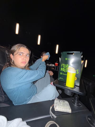 The image captures an interior scene within a darkened Cinemex movie theater, located in La Purísima, Mexico. In the foreground, a female individual with medium-brown hair, wearing a teal long-sleeved shirt and light gray pants, is seated in a black cushioned theater seat. She is observed looking to her right with a wide-eyed, grimacing facial expression. To her immediate right, a small black tray table holds a large, dark green "Cinemex" branded paper bag with "#TEAMPALOMOS" text and a bright yellow beverage container with a translucent lid positioned within a cup holder. Behind and to the right of the female, a male individual, also wearing a blue long-sleeved shirt, is partially visible. He is actively holding a light blue cloth with both hands, appearing to wipe a surface or object. A red cup is visible on a surface in front of him. The theater environment is characterized by dark walls and ceiling, with several vertical rectangular lights providing ambient illumination. A white, textured wallet or small clutch bag with a strap is visible on the armrest in the lower right foreground, and a light gray fabric item is partially seen on the seat cushion in the lower left.