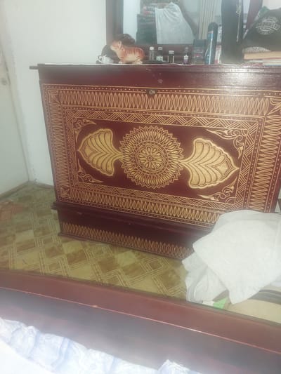 A dark red wooden chest with intricate gold geometric patterns and a floral design is positioned on a tiled floor. A tiger figurine, white towels, and other various objects are on top of the chest, suggesting an indoor setting. The scene is dimly lit, creating shadows and reflections on the surfaces. The location is identified as Ması, Kyrgyzstan.