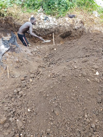 The image depicts a man digging in a dirt pit, possibly for mining or construction. The man is shirtless and wearing dark pants with red and white stripes. He is using a shovel to move dirt, which is flying through the air. In the background, there is lush green vegetation and dry grasses. A burnt tree stump is visible to the left of the man. The ground is covered in dirt and small rocks. The scene appears to be outdoors, likely in a rural or undeveloped area. The lighting suggests it is daytime, but the weather cannot be determined. There is no visible text in the image. The man's expression is focused and determined, suggesting he is engaged in strenuous labor. The overall impression is one of hard work in a natural environment.