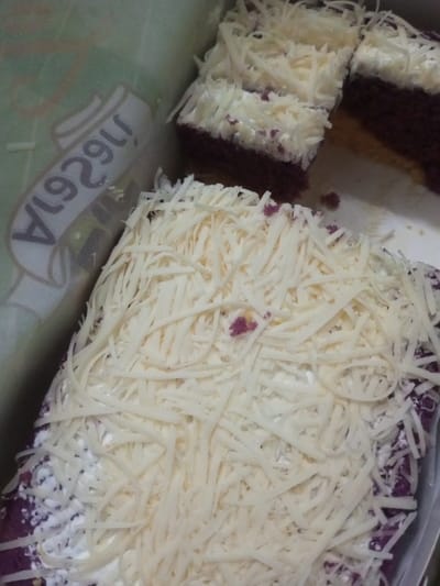 A close-up shot reveals slices of ube cake inside a container. The cake has a vibrant purple hue and is topped with a generous layer of white frosting and shredded cheese. One slice shows a layer of yellow cake between the purple base and the white frosting. Part of the container's logo is visible, featuring a word in white font.