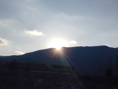 The image displays a mountainous landscape in Qoraqiya, Uzbekistan, during either sunrise or sunset. A brilliant sunburst, partially obscured by a dark, silhouetted mountain range, is centered in the upper middle and slightly to the right, emitting distinct light rays. The sky above is light blue with scattered white and light grey clouds. In the lower foreground, a building with a prominent reddish-brown, sloped roof is visible on the left, rendered in deep silhouette. To the right of this structure, another smaller, dark building or shed is also silhouetted. Dark, conical trees are present in the foreground, with one near the left building and another further to the right. The ground in the immediate foreground is dark, uneven, and appears to be rocky or covered with sparse vegetation. No individuals or active interactions are visible within the frame.
