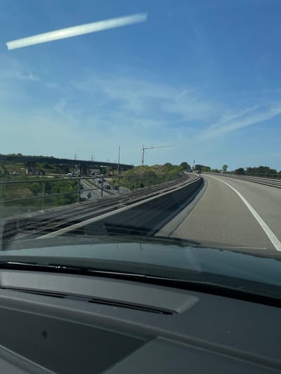 The image captures a view from inside a vehicle traveling on a highway, likely in or near Sankt Augustin, Germany. The highway curves to the right, with a white line marking the edge of the lane. To the left of the highway, there are train tracks running parallel, bordered by a metal fence and lush green vegetation. Beyond the train tracks and fence, a multi-lane highway is visible, carrying a moderate amount of traffic, including cars and trucks. In the distance, an elevated highway structure is under construction, supported by concrete pillars. Power lines and pylons are also present in the background. A construction crane is visible on the right side of the frame, further indicating ongoing infrastructure development. The sky is a clear blue with a few wispy clouds and a bright, elongated reflection, possibly from the sun or another light source, streaks across the upper portion of the image. The dashboard of the vehicle is visible in the foreground, suggesting the perspective of a driver or passenger.