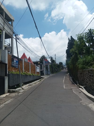 The image depicts a street in a residential area. The road is asphalt, and slightly uphill. On the left side, there's a building with visible signage including "24" and "GADOG," suggesting a commercial establishment. A wooden fence with red triangular features is also visible. On the right, a stone wall borders the street, with lush green foliage growing above it. Overhead, power lines crisscross the sky, which is partly cloudy with blue patches. In the distance, the road continues uphill, with glimpses of buildings and trees further ahead, and vehicles in the distance.