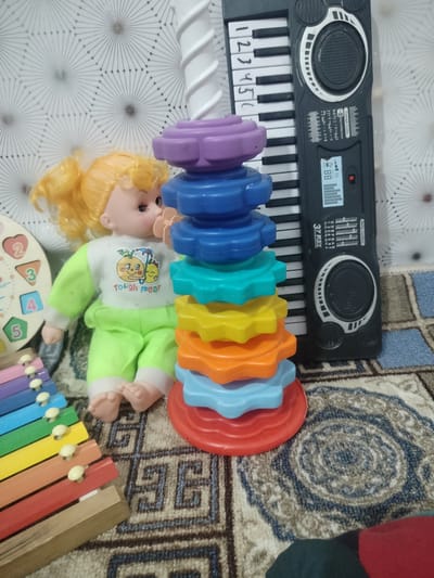 The image shows a collection of children's toys on a patterned rug. In the foreground, a colorful xylophone is visible. To the right of the xylophone, a doll with blonde hair is seated, wearing a light green outfit. A stack of brightly colored stacking rings is placed next to the doll. Behind the stacking rings, a toy keyboard with black and white keys is positioned. The background features a textured, possibly wallpapered, surface with a geometric starburst pattern. The overall setting appears to be indoors, likely a child's play area. The location in the city of Cholpon, Kyrgyzstan, is not determinable from the visual information presented.