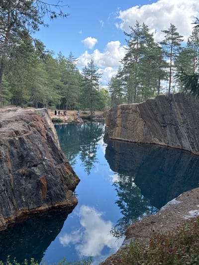 In Säter, Sweden, this image captures a serene quarry lake, a popular spot for swimming and diving, surrounded by a dense forest of pine trees. The deep blue water reflects the sky and the surrounding cliffs, creating a mirror-like effect. A group of people are gathered near the edge of the quarry, some appearing to be preparing for a dive, while others stand watching. The rugged cliffs, marked by layers of rock and sparse vegetation, rise sharply from the water's edge, hinting at the quarry's industrial past. The scene is bathed in the soft light of a partially cloudy day, with the sun intermittently breaking through the clouds, casting a dappled pattern on the water and surrounding landscape.