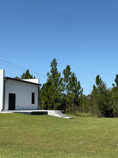 A modern white house with black trim and dark doors sits on a lush green lawn under a clear blue sky. The house has a simple, rectangular design with a flat roof and a few windows. A set of concrete steps leads up to a raised patio area in front of the house. Behind the house, a dense line of tall pine trees and other greenery forms a natural backdrop. Power lines stretch across the sky above the trees. The scene appears to be in a semi-rural or suburban area of Lehigh Acres, Florida, United States of America, characterized by open space, well-maintained landscaping, and a clear, sunny climate. There are no people or animals visible in the image.