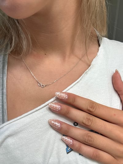 The scene, located in Rocklin, United States, features the upper torso and right hand of an individual, characterized by fair skin and light-colored hair. The subject wears a white, short-sleeved t-shirt, with a fragment of a gray garment visible on the left. A silver-colored chain necklace, adorned with a small infinity symbol pendant, is centrally positioned on the lower neck. The right hand, displaying almond-shaped fingernails coated in a translucent light pink or nude polish with distinct white polka dots, rests against the upper chest. The immediate background is an unidentifiable, plain, light-toned surface, consistent with an indoor environment.