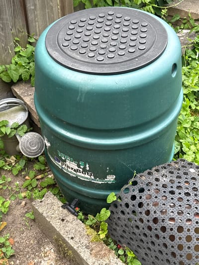 A green water butt, also known as a rainwater harvesting barrel, is situated outdoors. It has a textured black lid with numerous circular indentations. The barrel itself has ribbed detailing around its circumference and a label that reads "Water Butt" along with other illegible text. To the left of the water butt are two galvanized metal watering cans. A black perforated mat is placed in front of the water butt. The scene is surrounded by green foliage and a wooden fence is visible in the background. A concrete block is in the foreground, with a small red tap protruding from it. The overall environment suggests a garden setting. This image was taken in Bemmel, Netherlands.