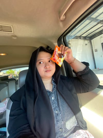 A woman wearing a black hijab and a denim jacket is inside a vehicle, holding a chocolate bar wrapper in front of her eye. The vehicle appears to be parked or moving slowly, as indicated by the background view of houses and vegetation. The sunlight streams in through the car window, illuminating the scene. The woman's expression is playful as she looks away from the camera. The interior of the car is visible, with beige upholstery and air vents. The location is specified as Randubango, Indonesia.