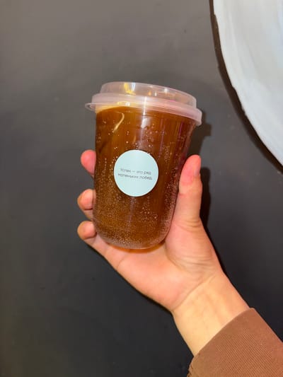 A hand holds a transparent plastic cup filled with a dark brown beverage, likely coffee or iced tea, with visible bubbles. A small, round, light blue sticker with white text in Russian is affixed to the cup. The text translates to "Success is a series of small victories." The background is dark gray. The image is a close-up shot, suggesting it was taken indoors, possibly in a home or cafe environment. The presence of the cup and hand indicates a person is interacting with the beverage. The location in Veshki, Russia, is not discernible from the visual elements of the image itself.