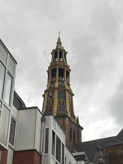 The image shows a tall, ornate clock tower, likely a church tower, in Groningen, Netherlands. The tower is predominantly gold and dark gray, with visible clock faces and brickwork. In the foreground, to the left, a modern building with large windows and brick base is partially visible. The sky is overcast and cloudy.