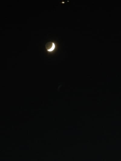 The image is a dark, nighttime photograph of the sky over Algés, Portugal, featuring a crescent moon. The moon is illuminated from the right, displaying a thin sliver of light against the black expanse of the night sky. A faint, darker shape is visible below and to the right of the moon, possibly a shadow or atmospheric distortion. In the upper portion of the image, a cluster of small, bright lights are faintly visible, potentially distant city lights or other celestial bodies. The overall scene is stark and minimalist, focusing on the celestial elements against the backdrop of darkness.