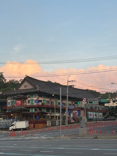 In the city of Pocheon-si, South Korea, an image captures a street scene at dusk. The sky is a soft blue with large, fluffy pink clouds, indicating the sun is setting. A traditional Korean building with a dark tiled roof dominates the mid-ground, its facade adorned with green shutters and balconies. Red and blue lights are visible along the balconies. In front of the building, a white delivery truck is parked on the side of the road. The street is marked with crosswalks and lined with orange traffic cones. Traffic lights are visible, with green lights illuminated. A street sign indicates "Chukseok Jct" and the Korean text "축석고개로". The overall atmosphere is calm and serene, with the warm colors of the sky reflecting on the clouds.