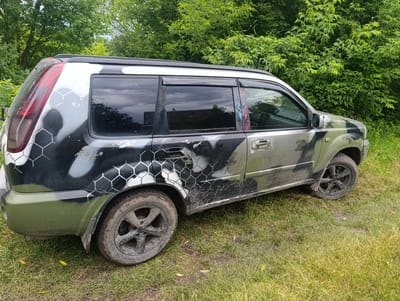 A utilitarian, slightly weathered dark gray Nissan X-Trail SUV, customized with a distinctive camouflage paint job featuring a hexagonal pattern, is parked on a patch of overgrown grass and dirt. The SUV is angled towards the right, showcasing its side profile. The camouflage paint is a mix of black, gray, and olive green, with some areas appearing worn or chipped, revealing the original paint underneath. The vehicle is situated outdoors, with a dense backdrop of lush green trees and foliage, suggesting a rural or semi-rural setting. The sky is overcast, reflecting in the tinted windows of the car. The tires are dirty, indicating recent use on unpaved surfaces. The scene appears to be static, with no people or ongoing activities evident. This image was captured on June 17, 2023, at approximately 10:05 AM in Novoselytsya, Ukraine.