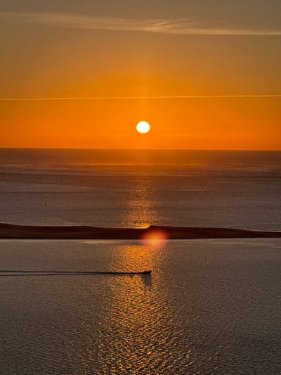 The image depicts a serene sunset over the Arcachon Bay in La Teste-de-Buch, France. The sun, a bright orb, hangs low on the horizon, casting a warm, orange glow across the sky and water. A thin contrail stretches across the upper portion of the sky, a subtle indication of human activity.
The water itself is a gradient of deep blues and grays, reflecting the fiery hues of the sunset. A long, narrow strip of land, likely the Dune of Pilat, forms a dark silhouette across the middle ground, separating the bay into two distinct bodies of water.
In the foreground, a small boat is making its way across the water, leaving a subtle wake behind it. The boat is silhouetted against the shimmering reflection of the sun on the water, creating a dynamic contrast. The scene is one of tranquility and natural beauty, capturing the essence of a peaceful evening by the coast.