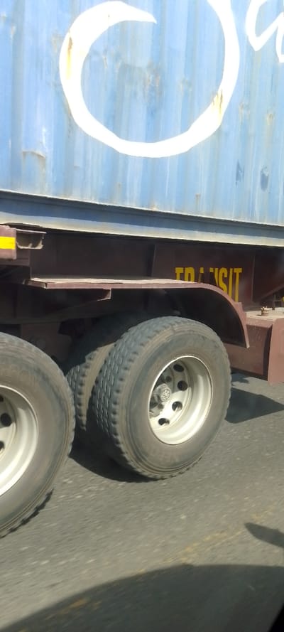 The image depicts the side of a large blue shipping container secured on a maroon trailer chassis, traveling on an asphalt road in Kinanie ward, Kenya. The container's corrugated metal surface is weathered, displaying areas of rust and faded light blue paint. A large, thick white marking, resembling a partial circle or the letter "C," is visible on the upper section of the container. Below the container, the dark maroon chassis features yellow stencil lettering, partially visible, spelling "TRANSIT." Three large black truck tires are visible, two forming a dual-wheel axle and one from an 앞/rearward axle, all with silver multi-hole rims. One rim's hubcap displays a logo that appears to read "DBP." The road surface beneath the tires is dark asphalt, suggesting a clear day with a strong shadow from an unseen object or vehicle cast onto the road in the lower right foreground. No individuals are visible. The scene implies motion, likely captured from a parallel moving vehicle.