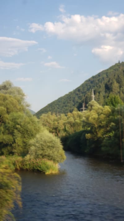 The image is a slightly blurry, vertical shot of a river flowing through a valley in Švošov, Slovakia. Lush green trees and foliage line the banks of the river, with a steep, densely wooded hillside dominating the background. The sky above is blue with scattered white clouds. In the distance, on the hillside, a transmission tower is visible. The river itself is dark and appears to be flowing calmly. The overall scene depicts a natural, tranquil landscape.