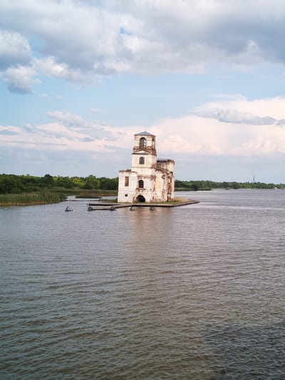 In the city of Krokhino, Russia, an old, white, decaying church stands on a small island in the middle of a wide, calm body of water. The church is multi-story with a bell tower. Its walls are weathered and crumbling, exposing brickwork in some areas. Multiple windows, some arched and others rectangular, are visible, many of them appearing empty or broken. The building appears to be in a state of disrepair.
The water surrounding the island is a murky brown and has a rippled surface. Along the shore of the island, wooden structures resembling docks or platforms are visible, some appearing partially submerged. In the water closer to the shore, there are several dark, angular objects, possibly remnants of submerged structures or debris.
To the left of the church, tall reeds grow along the edge of the water, forming a dense barrier. Beyond the reeds and water, a thick line of green trees stretches across the horizon, forming a lush, forested background. The sky above is partly cloudy, with large, white cumulus clouds against a pale blue sky. The lighting suggests it is daytime, possibly late afternoon given the softer light. The scene is tranquil and presents a solitary, weathered structure against a natural backdrop.