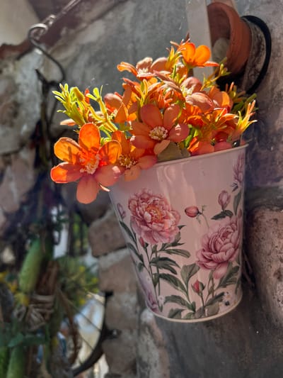 A close-up shot captures a decorative floral arrangement in a pink, patterned pot hanging on a rough, textured stone wall. The pot, adorned with pink roses and green leaves, holds a vibrant bunch of artificial orange flowers and green foliage. Sunlight streams in from the right, illuminating the flowers and casting shadows on the wall. In the background, hints of greenery and a dark circular object are visible, suggesting an outdoor setting, potentially in the city of Belo Pole, Bulgaria, where such decorative arrangements are common in residential courtyards or on balconies. The scene is static, with no people or animals present. The focus is on the pot and its contents, with the backdrop providing context.