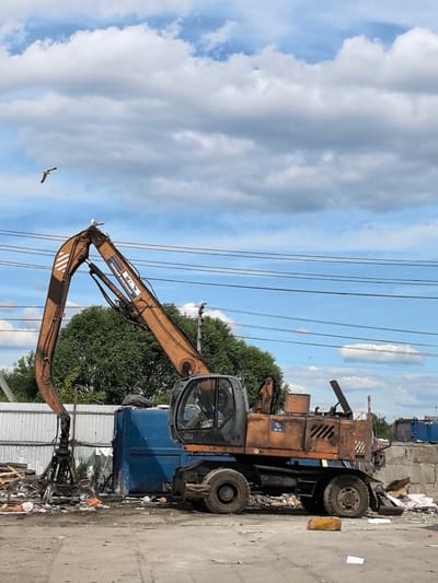 An orange excavator with a grapple attachment is positioned in an outdoor industrial area, possibly a scrap yard or recycling facility, in Novo-Talitsy, Russia. The excavator is situated on a dirt and gravel surface, with various piles of debris and scrap metal scattered around it. To the left of the excavator, there is a tall, corrugated metal fence, and to the right, a concrete wall. In the background, there are trees and other industrial structures, including large blue bins. Several power lines are visible overhead against a cloudy blue sky. A seagull is perched on the excavator's boom, and another is in flight in the sky. The scene appears to be a functional industrial site during daytime.