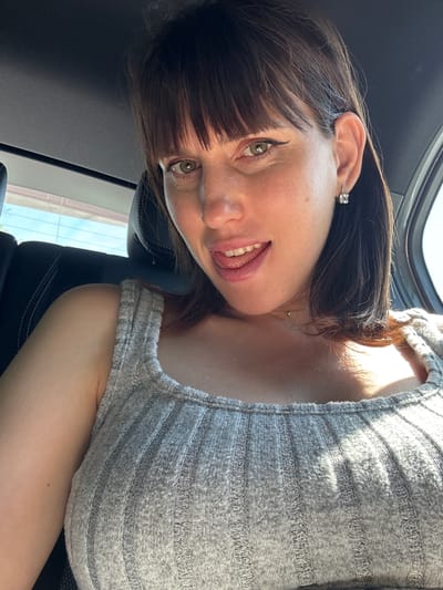 A woman with dark hair and light skin is photographed from a close-up, low-angle perspective inside a vehicle. She is wearing a gray striped top and an earring. The interior of the vehicle is visible with a black headrest and part of a window. The location is not identifiable in the city of Pomezia, Italy from the available information.