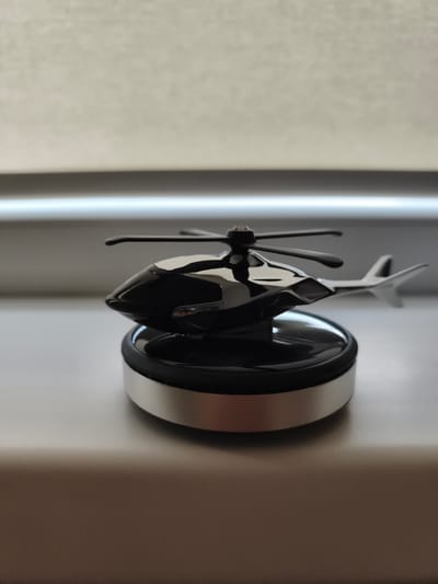 This media file contains an image of a shiny black helicopter figurine placed on a metallic base. The figurine has a sleek, modern design with a prominent rotor and tail fin. It is positioned on a smooth, light-colored surface, possibly a windowsill or desk, with a blurred background that suggests an indoor setting. The lighting is soft, casting gentle reflections on the metallic surfaces of the helicopter and its base. The overall impression is one of a decorative item, possibly a desk ornament or a collectible. There is no visible text in the image. The setting appears to be in Dobele, Latvia, though no specific landmarks or indicators confirm this within the frame. The time of day is indeterminate due to the controlled indoor lighting. The image focuses solely on the object, with no people or activity present.