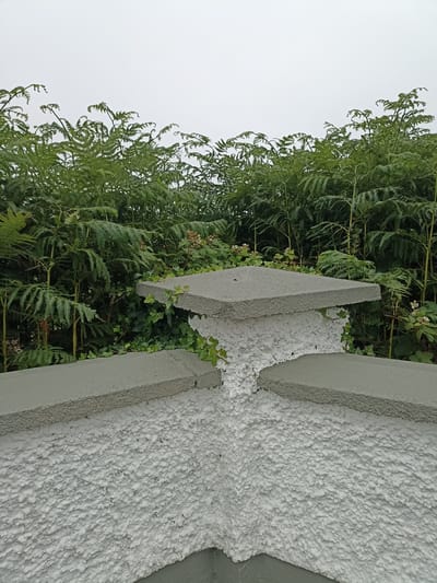 This image depicts a close-up view of the top of a textured white concrete pillar in Annagry West, Ireland. The pillar is capped with a flat, grey concrete slab. Two grey concrete ledges extend outwards from the top of the pillar. Behind the pillar, a dense growth of green bracken ferns and some ivy are visible. The sky is overcast and appears pale grey. There are no people or animals in the image.