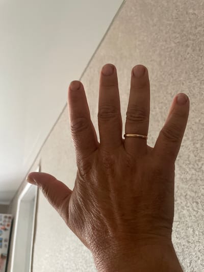 A person's hand with a thin gold ring on the ring finger is raised in front of a light-colored, textured wall. The hand is positioned to face the camera, with the fingers spread slightly apart. The lighting appears to be natural, casting soft shadows. The background consists of a wall with a textured pattern, possibly wallpaper or a similar material.