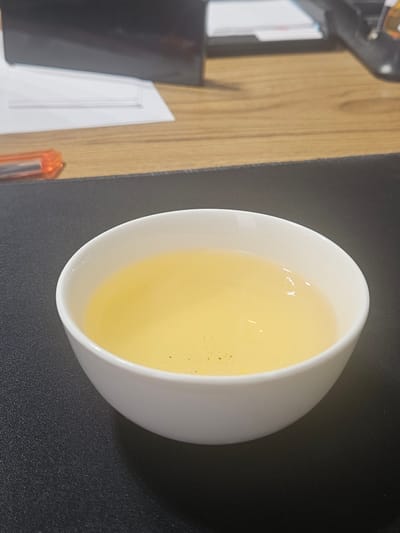 The image shows a white ceramic cup filled with a pale yellow liquid, likely tea, on a dark gray textured surface. The cup is positioned slightly off-center, towards the bottom right of the frame. In the background, a wooden surface is visible, with a stack of papers and a portion of what appears to be a black electronic device. An orange pen or marker is also partially visible on the wooden surface. The overall scene suggests an indoor setting, possibly an office or a desk area. There are no people or animals present in the image. The lighting suggests it is daytime, with some reflections visible on the surface of the liquid in the cup. The location in the city of Uchtepa, Uzbekistan, is not discernible from the visual information provided in the image.