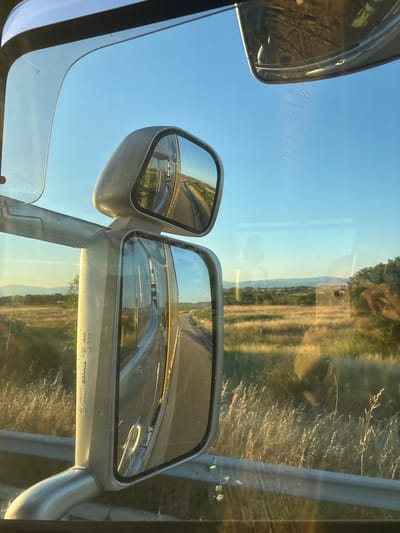 The image is a first-person perspective from inside a vehicle, looking out through the side window. It is a sunny day with a clear blue sky. The foreground features two side-view mirrors of the vehicle, with a metallic arm extending between them. The mirrors reflect the road ahead, which appears to be a highway or a long, straight road winding through a rural landscape.
The landscape outside the window consists of dry, golden grass and scattered trees. In the distance, a range of low mountains is visible under the bright sky. The lighting suggests it is either early morning or late afternoon, with the sun casting long shadows and illuminating the dry vegetation with a warm glow.
The image is taken in Aetokoryfi, Greece.